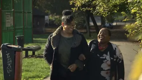Pat Simoyi and Dee Moore laughing as they walk arm-in-arm in a park