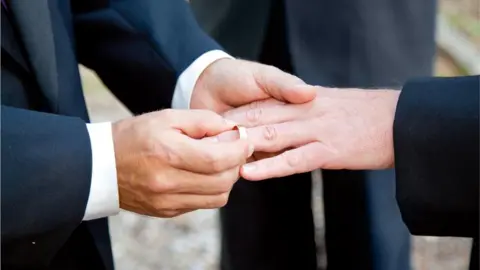 Getty Images One groom placing a wedding ring on another groom's finger