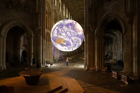 Oli Scarff / AFP A member of staff stands beneath artist Luke Jerram's installation Gaia, a replica of planet Earth, suspended in the nave of Ely Cathedral in Cambridgeshire