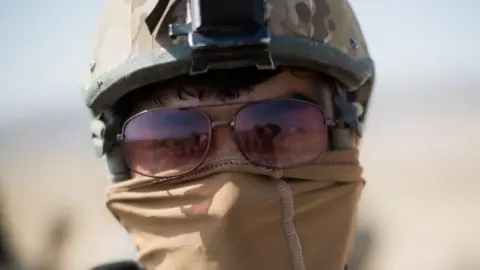 Benjamin Taggart A members of the Triples pictured front on with his head in shot. He is wearing a combat helmet, sunglasses, and his mouth is covered