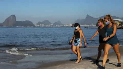 Getty Images People walk wearing a protective mask on Icarai beach, as the Sugarloaf Mountain is seen in the distance, during the coronavirus (COVID-19) pandemic on May 21, 2020 in Niteroi, Brazil