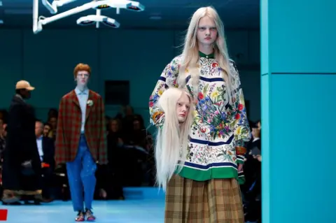 Reuters Female model in floral dress walks down the catwalk holding a mannequin head that is a replica of her head