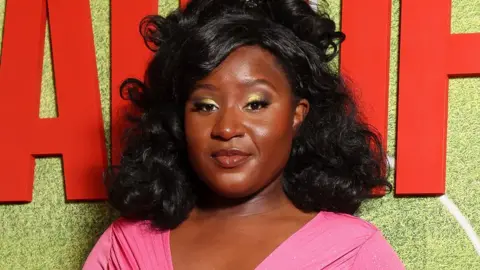 Getty Images Susan Wokoma. Susan is a black woman in her 20s and is pictured at a red carpet event. Susan has shoulder-length curly black hair which is styled half-up-half-down. She has brown eyes and wears a gold eyeshadow and eye-liner make-up look. She wears a low-cut bubble gum pink dress and smiles at the camera