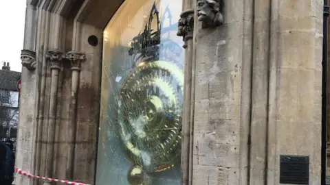 Orla Moore/BBC Red and white tape cordons off the protective glass on the side of a building. The smashed and cracked glass can be seen and behind it you can see the circular gold clock with a grasshopper sculpture at the top.