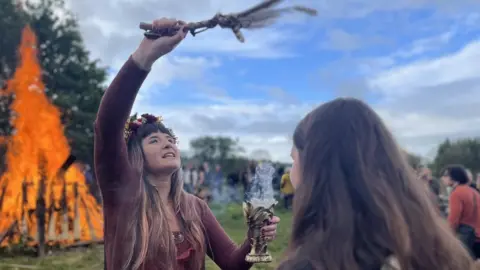 Jasmine Ketibuah-Foley/BBC A woman waves wood above another woman's head, she is wearing a headband and a large fire is burning in the background