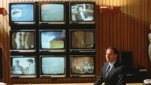 Getty Images A younger Silvio Berlusconi sits in an ornate wood-panelled office with nine very large (for 1986) televisions arrayed in a grid on the wall behind him