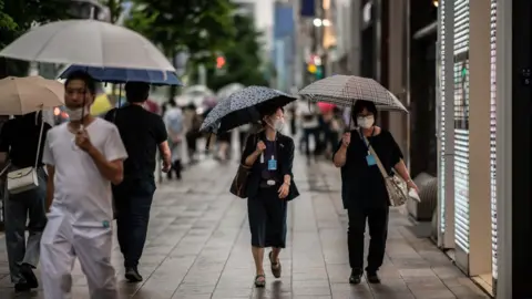 Getty Images People wearing face masks shelter from the rain under umbrellas as they walk along a street on 30 June, 2020 in Tokyo, Japan.