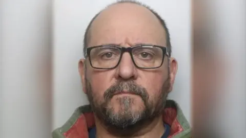 Northamptonshire Police Man with very short brown hair, a beard and glasses wearing a green and yellow coat