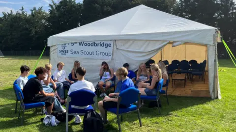 Shaun Whitmore/BBC Sixth form students seated outside a tent. The tent has a sign for the 5th Woodbridge Sea Scouts.