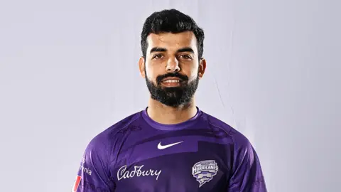 Getty Images Shadab Khan against a white studio background. He's got short hair and a trimmed but thick-looking dark beard. He's smiling and wearing a purple t-shirt with a Nike "swoosh" logo over the upper chest. Below that on the camera left is an embroidered Cadbury logo - in cursive writing and white text. On the right is the Hobart Hurricanes' team logo - the word Hurricanes in angular white text over a grey, precisely-drawn shaped made up of curved rectangles that decrease in size from top to bottom to create the effect of a hurricane or funnel cloud.