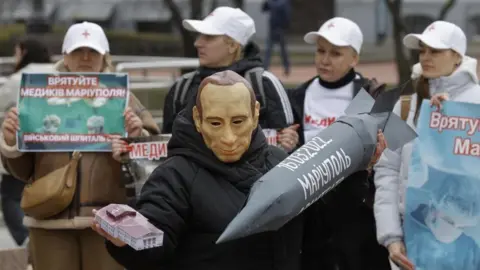 SERGEY DOLZHENKO/EPA-EFE/REX/Shutterstock A man wearing a mask depicting Russian President Vladimir Putin commemorates the second anniversary of the shelling of the Mariupol Drama Theatre where more 300 civilians were killed