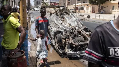 Getty Images People walk past a burned out car in Dakar, on June 5, 2023, as protest calmed down four days after a court in Senegal sentenced opposition leader Ousmane Sonko, a candidate in the 2024 presidential election, to two years in prison on charges of "corrupting youth" but acquitted him of rape and issuing death threats