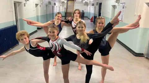 BBC Six young gymnasts holding their legs up in a prison corridor