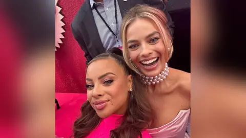 Danica Taylor Danica Taylor and Margot Robbie posing for a selfie at the Barbier premiere, Margot looks very happy and Danica is smiling. Margot is wearing a pearl necklace and has her blonde hair tied up while Danica has her brunette hair worn down. Both are wearing pink dresses