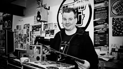 Clare Butchart Tom Butchart, holding a vinyl record, behind the counter at Sound It Out Records