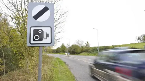 Ben Birchall/PA Media National speed limit sign with car (distorted by motion blur) speeds by on country road