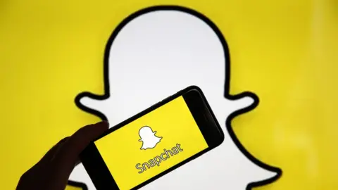 Getty Images The Snapchat ghost logo is seen as a background with a phone and the app held up in front of it