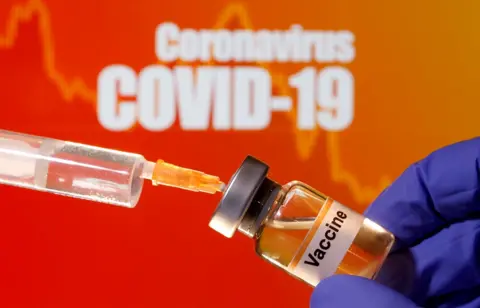 Reuters A small bottle labelled with a "Vaccine" sticker is held near a medical syringe in front of displayed "Coronavirus COVID-19" words in this illustration taken