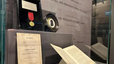 A glass case holding a the medal, which has a red ribbon, mounted on a black background, along with a pamphlet outlining the case for the creation of the service.