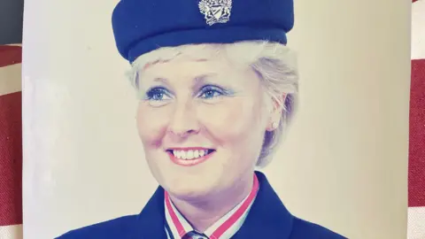 Handout Image of Anne in her cabin crew uniform - a navy jacket with a matching hat. She has light blue eye shadow and red lipstick on. 
