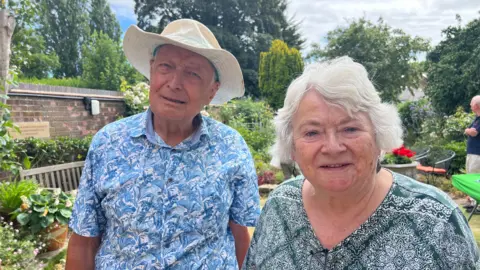 A couple looking directly into the camera. They are stood infront of a garden. An older woman is on the right. She has short white hair and is wearing a green shirt. A man is stood to the left of her wearing a white hat and a blue floral shirt.