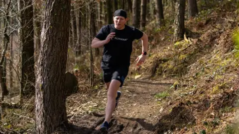 PA Media A man in a black hat, t-shirt and shorts is seen running through woodlands. 