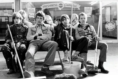Ella Murtha A black-and-white photo of four young men sitting in what looks like a shopping centre and looking into the camera with straight faces. They are holding various mobs and brooms and are dressed in casual clothes.