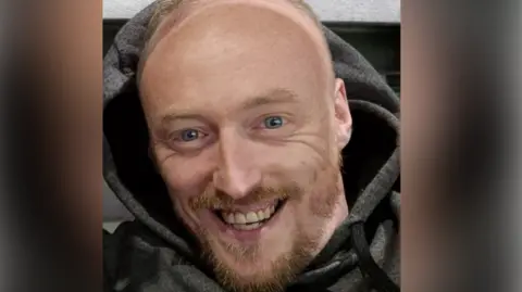 A man smiles as he takes a selfie. He is bald and has a ginger beard. He is wearing a dark grey hooded top.