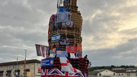A large unlit bonfire covered in British and Northern Ireland flags beside a road. 