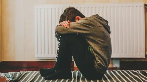 Getty Images An upset-looking young boy wearing a grey hoodie sits hunched over on the floor with his head tucked into his arms, which are folded on top of his raised knees