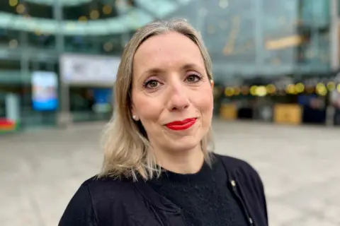 Jo Thewlis/BBC Sarah Taylor is wearing bright red lipstick and a black top and cardigan. She is standing in front of the glass-fronted Forum building in Norwich.