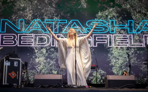 Paul Campbell Natasha Bedingfield wearing a white flowing gown holds out her arm while performing on stage 