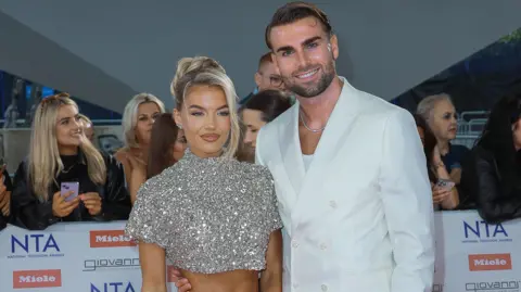 Getty Images Molly Smith and Tom Clare post together on a red carpet. Molly is wearing a sparkly cropped top and Tom is wearing a white suit over a white top.  