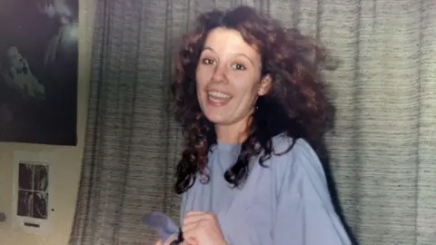 A picture taken in the 90s showing a girl in her 20s smiling at the camera, she is wearing a baggy t-shirt and has curly hair with flashes of red. She's in front of a curtain and you can see pictures on the walls to the right, it gives the impression it is taken in a military dorm room.