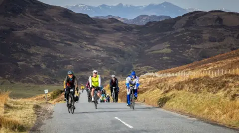 Paul Campbell A group of riders pedal up a hill on a rural road. Two cyclists, on the right, are riding a tandem and the cyclist at the front is wearing a Batman costume. Behind the cyclists rise steep hills and mountains.