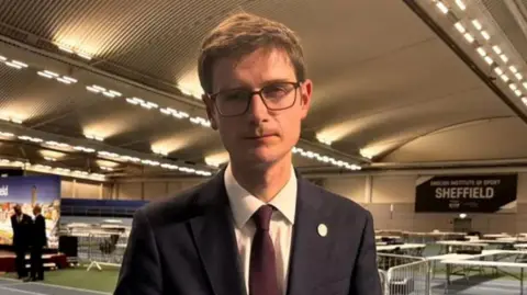 BBC/Simon Thake Tom Hunt: a man with glasses, wearing a dark jacket, white shirt and burgundy tie, pictured at the Sheffield election count