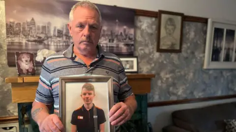A man with grey hair and a grey polo top holds up a picture of a young man. The photo of the older man is taken in his living room