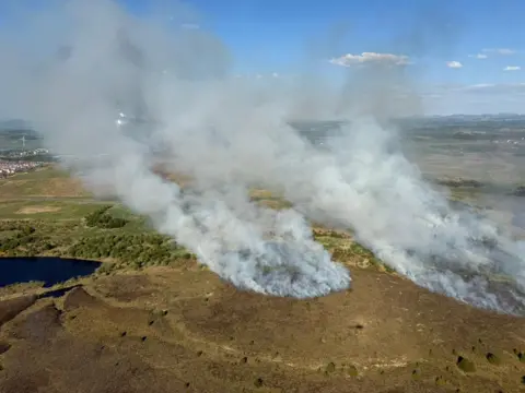 SFRS Grey smoke rises from a bracken-covered hill on a sunny day in a photo taken from a helicopter 