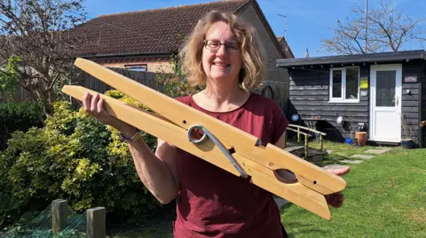 Cat Thompson has light brown hair and wears glasses, she is wearing a burgundy T-shirt and is holding a giant clothing peg which is about two feet (0.61m) long. She is in a sunny back garden, with a black shiplapped garden shed behind her on a grass lawn.