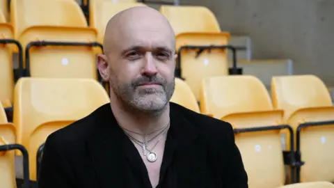 Shaun Whitmore/BBC Andy Wall a man who is looking at the camera and sat on a yellow seat at Carrow Road Football stadium.