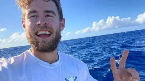 Jack Jarvis Jack Jarvis at sea wearing a t-shirt and smiling at the camera while doing a "peace" or "v for victory" sign with his hand.