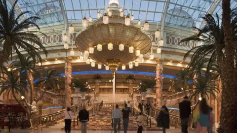 Trafford Centre A large chandelier hangs over a grand staircase in a shopping centre, with shoppers walking beneath it and palm trees all around.