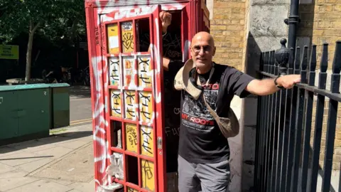 A man in a black t shirt and grey shorts stood next to a red phonebox