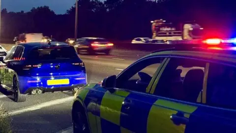 A blue car is parked on the side of a busy road and a police car is parked behind it. The image is taken in the dark and the lights from several other vehicles travelling along the road can be seen