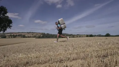 Photo of Firmstone running across a stubbly hay field, with the hay bale - wrapped in plastic sheeting - on his back. He is wearing a blue t-shirt and black shorts. The sky is blue and there is strong sunshine. 