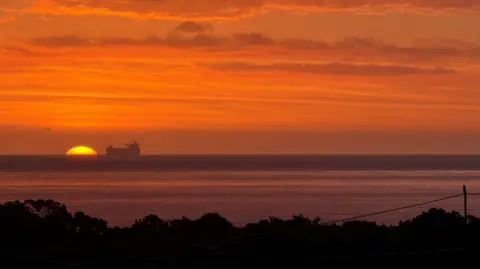 Brian Rodger An image of an orange sunrise sky, with the sun appearing over the North Sea horizon, next to an offshore vessel in the distance.