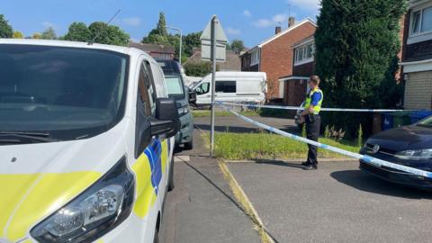 A police van parked near the curb on the street where the house is with a police officer behind a cordon of tape which is in front of the house's driveway