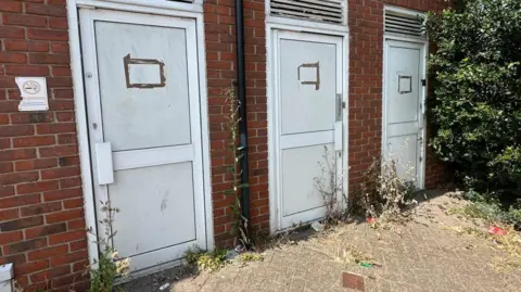 BBC/Charlotte Benton Three white plastic derelict doors, leading to three public toilets, with overgrown weeds in front of them.