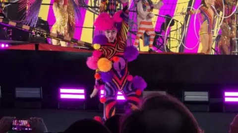 Ellis Grover Millie Harvey dressed in a fluorescent purple, pink, orange and black circus costume covered in pompoms on stage at the Take That concert.