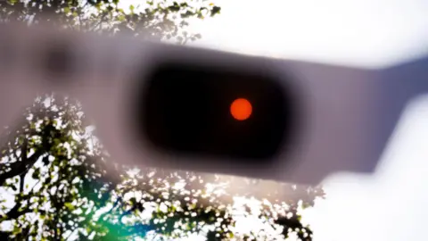 PA Media The sun polarised through a pair of Celestron solar eclipse glasses. There is a bright orange dot in one of the lenses of the glasses. The sky in the background is bright white and there is a leafy tree in the bottom left corner.